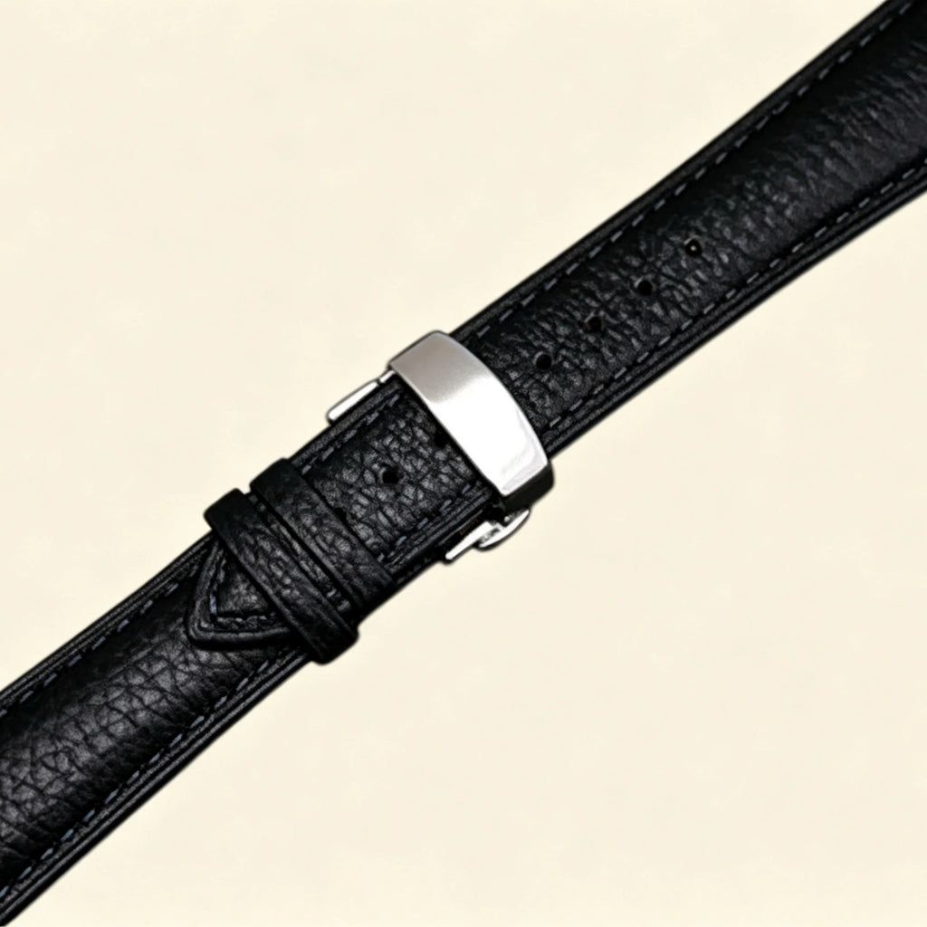 Black pebble-textured genuine leather Apple Watch strap — diagonal close-up showing stainless steel butterfly deployment clasp and strap taper, cream background