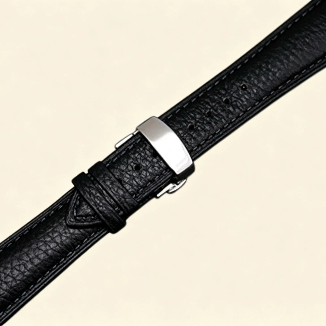 Black pebble-textured genuine leather Apple Watch strap — diagonal close-up showing stainless steel butterfly deployment clasp and strap taper, cream background