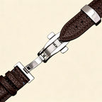 Dark espresso brown genuine leather Apple Watch strap — diagonal close-up showing stainless steel deployment clasp closed and cream topstitching detail, cream background