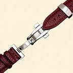 Burgundy pebble-grained genuine leather Apple Watch strap — diagonal close-up showing stainless steel deployment clasp closed and cream topstitching detail